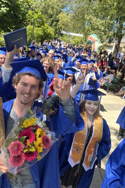 many graduating students hold up diplomas and flowers and smile at the camera as they walk out of the ceremony