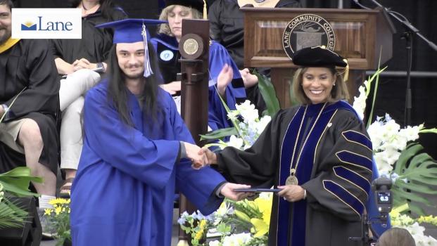 a tall young man in graduating blue cap and gown shakes hands with President Bulger as he accepts his diploma on stage