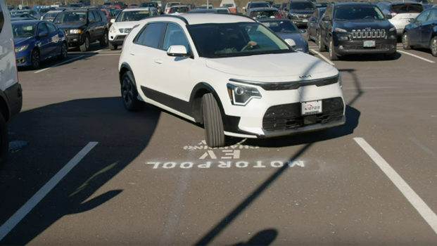 an electric vehicle, a white Kia sedan, pulls into a motorpool parking lot on a sunny day