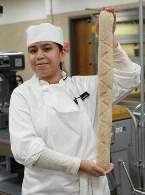 a culinary baking student in a chef uniform holding her baguette in a kitchen classroom